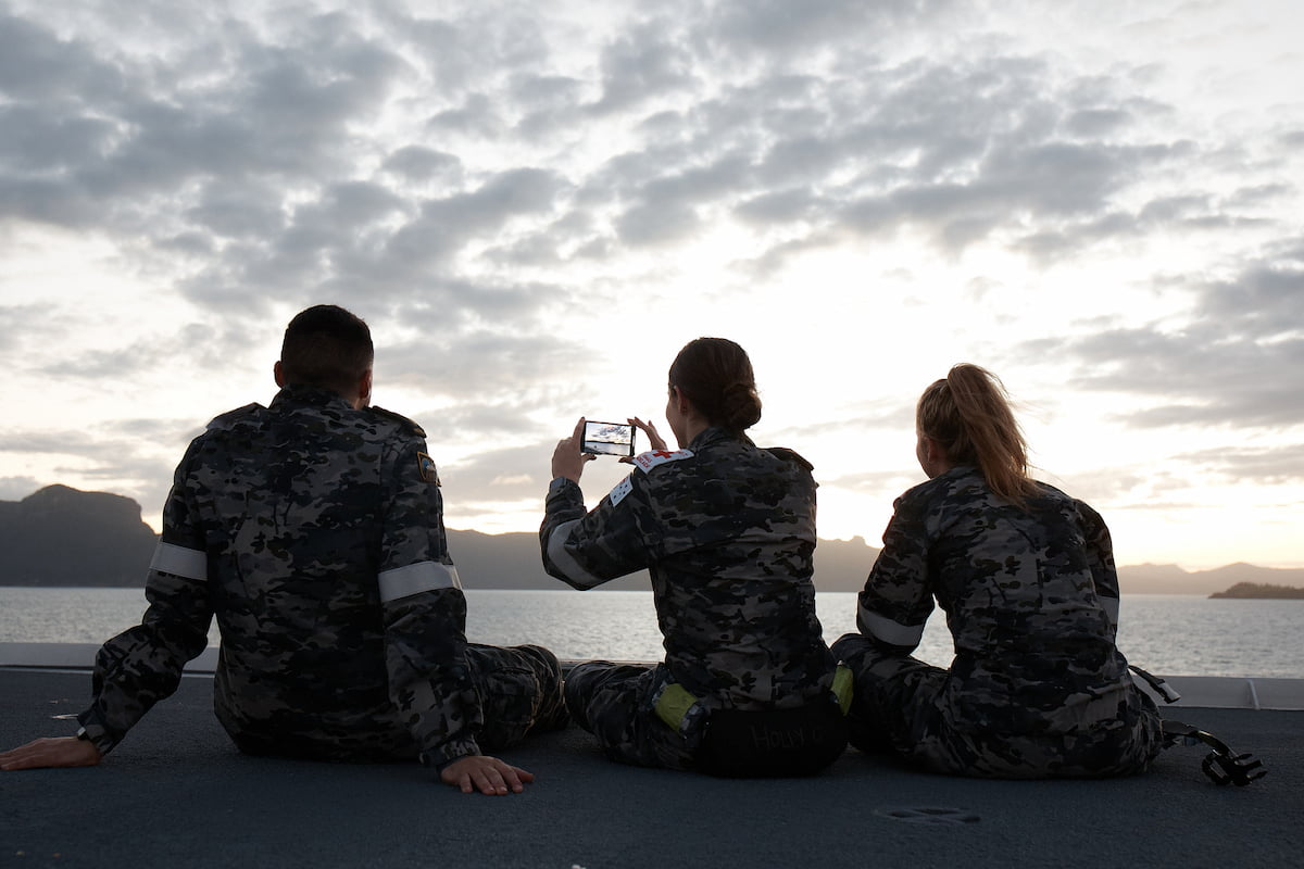 Three Navy personnel relax on the deck of a vessel at sea taking a photo of their surrounds.